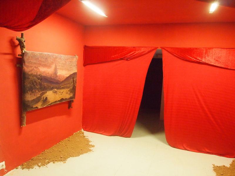 Daniel De la Barra, Instalación de paredes rojas con un cuadro de un paisaje montañoso enmarcado con troncos naturales y tierra en el suelo.