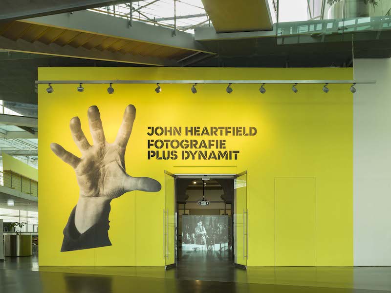 John Heartfield. Vista de la entrada a la exposición John Heartfield - Photographie plus Dynamit de color amarilla con una mano abierta.