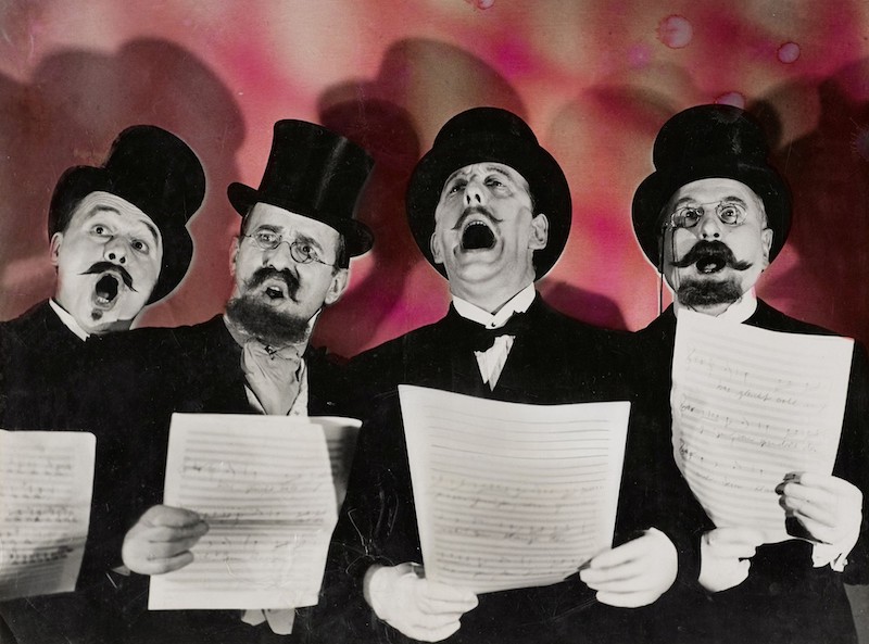 John_Heartfield, cartel para obra de teatro donde se ven 4 señores con sombreros de copa recitando con una roja de papel en la mano