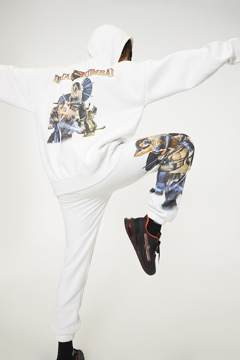 Pull and Bear x Mortal Kombat Chico con camiseta y pantalón de chándal blancos, con ilustraciones de Mortal Kombat de espaldas y con una pierna levantada.