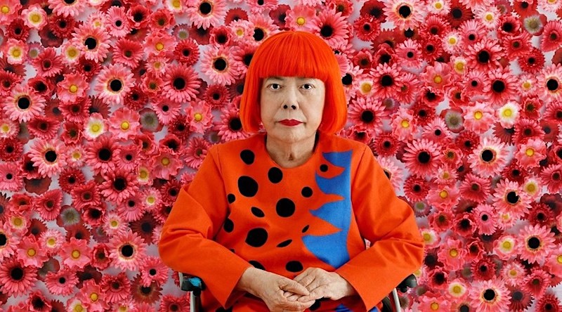 Yayoi Kusama, foto en de la artista con peluca roja delante de una pared de margaritas rojas