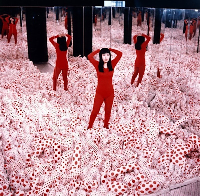 Yayoi Kusama, foto de la artista en una de sus instalaciones, una habitacion rodeada de espejos con muñecos de trapo de lunares blancos y rojos
