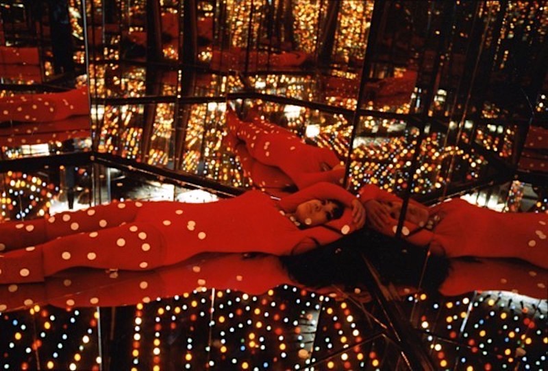 Yayoi Kusama la artista vestida de rojo tumbada en su instalacion llena de luces redondas rodeada de espejos y hexágonos en el suelo