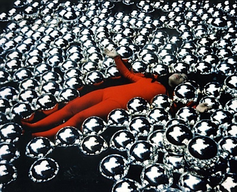 Yayoi Kusama la artista vestida de rojo tumbada en su instalacion llena de bolas de acero
