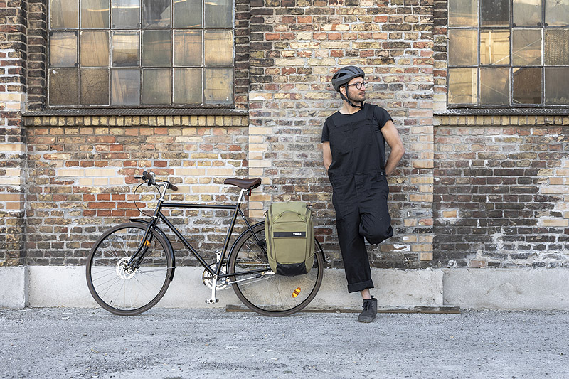 Thule_Paramount, foto de ciclista urmabo apoyado en una pared, a su lado hay una bici con alforjas
