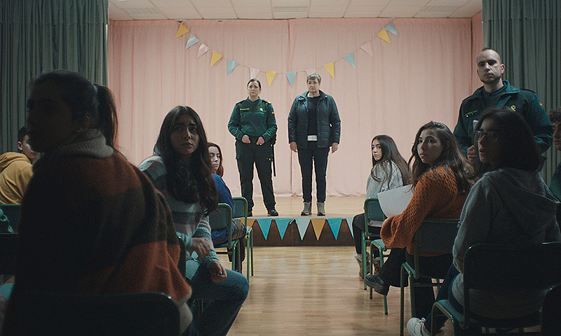 As Neves - fotograma de la película, se ve a una pareja de la Guardia Civil en el escenario de un salón de actos donde hay chicos y chicas