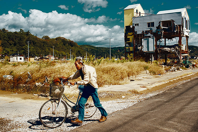 Jordi Socías. Al final de la escapada - un anciano en Fukushima empuja su bicicleta con edificio en ruinas al fondo