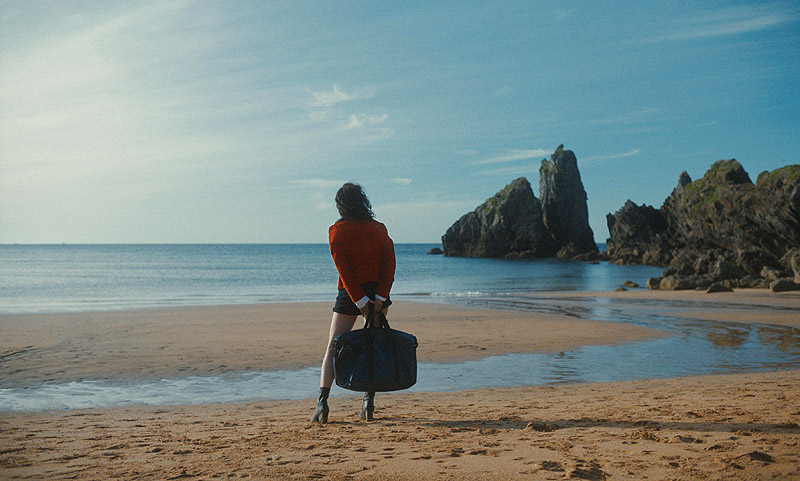 Nina, fotograma de la película, se ve a una mujer en la playa vestida de espaldas mirando al mar con una maleta en las manos