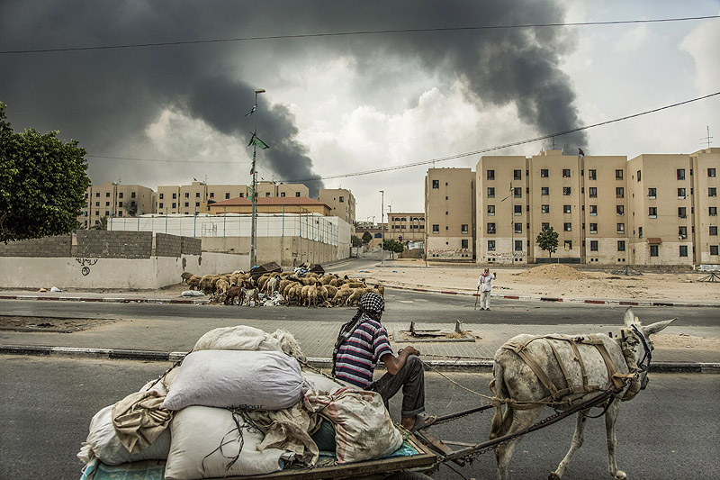 Impacte! - fotograma de documental, Una columna de humo se eleva hacia el cielo mientras dos hombres intercambian información en las calles vacías de Shiek Ziyad, en el norte de Gaza.