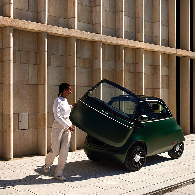 Microlino automóvil: Una chica abre la puerta de un microlino verde mate frente a un edificio