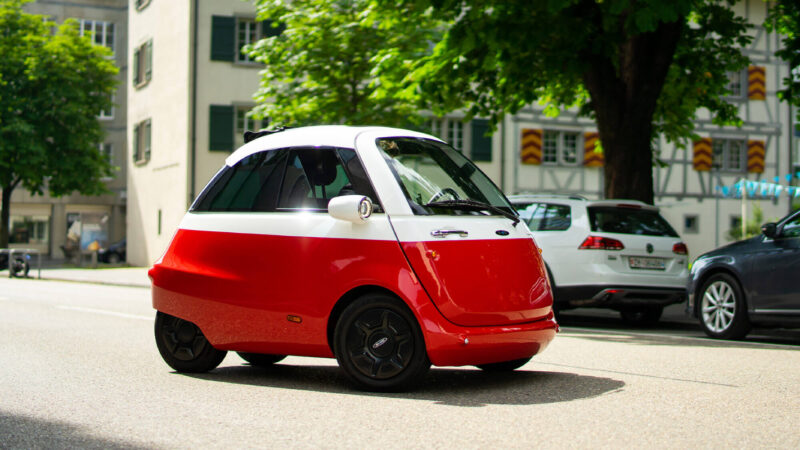 Microlino automóvil: Un Microlino rojo con el techo blanco por la ciudad rodeado de coches y edificios