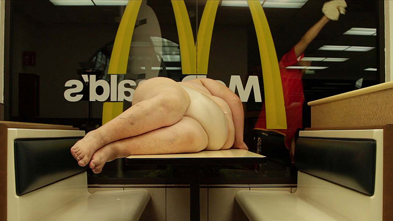 Festival Proyector - fotograma de video - una mujer obesa tumbada en una mesa de Mc Donald