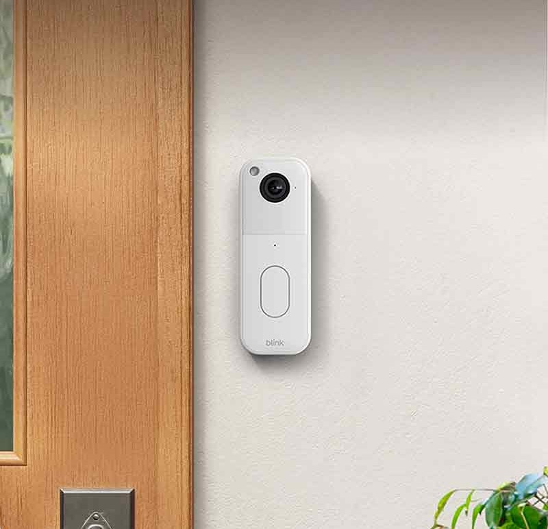 Blink Video Doorbell: dispositivo Blink Video Doorbell en blanco colocado en la pared de la entrada junto a una puerta de madera.
