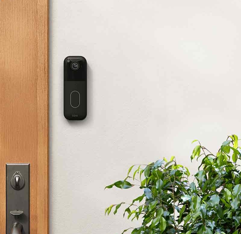 Blink Video Doorbell: dispositivo Blink Video Doorbell en negro colocado en la pared de la entrada junto a una puerta de madera.