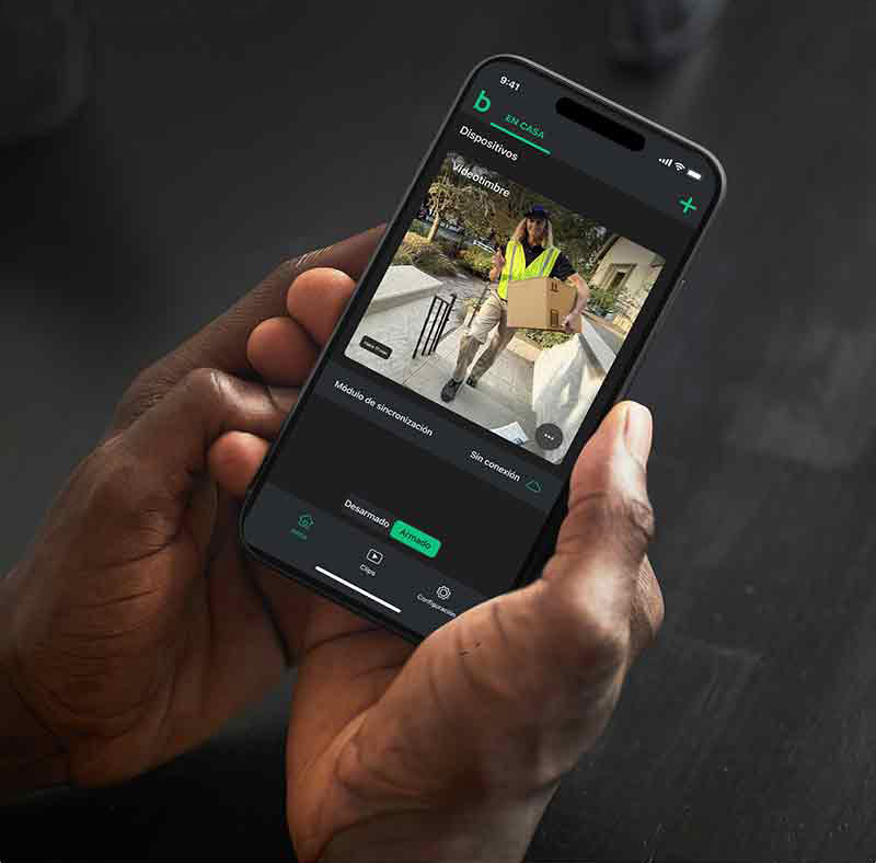 Blink Video Doorbell: las manos de un hombre de piel oscura sostienen un smartphone que muestra la imagen de la cámara de Blink Video Doorbell en la que aparece una mensajera que lleva un paquete.