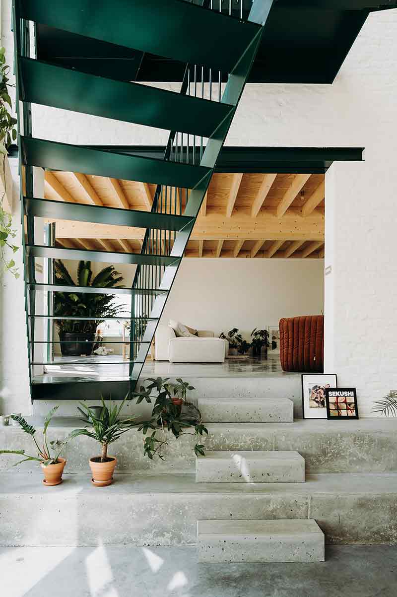 Escalera metálica interior de Casa Lyco, diseñada por Oyo Architects, sobre peldaños de hormigón y entre vegetación interior