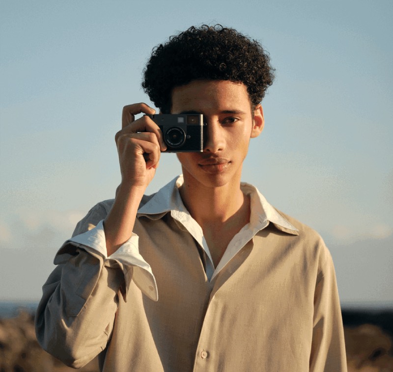 Fujifilm X half: retrato de un chico joven mulato de pelo afro que viste una camisa blanca y otra beige encima y mira por el visor de la cámara digital compacta Fujifilm X half en un paisaje natural un día soleado.