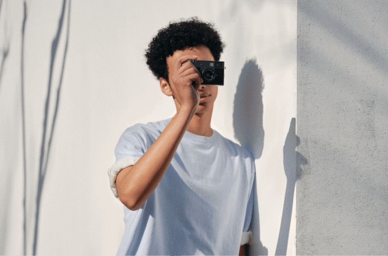 Fujifilm X half: retrato de un chico joven mulato de pelo afro que viste una camiseta azul claro y mira la pantalla de la cámara digital compacta Fujifilm X half mientras captura un momento de un día soleado.