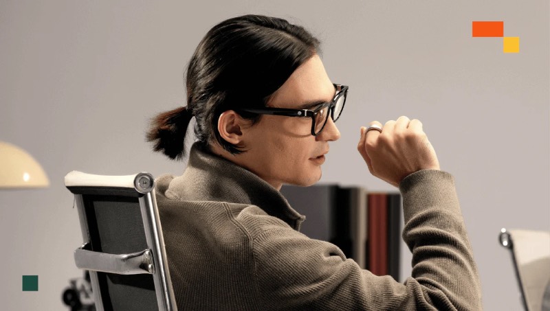 Halliday Glasses: un hombre con el pelo corto recogido en una coleta de lateral, sentado en una silla de oficina con un jersey en color tierra lleva puestas las Halliday Glasses y un anillo con el que las controla.