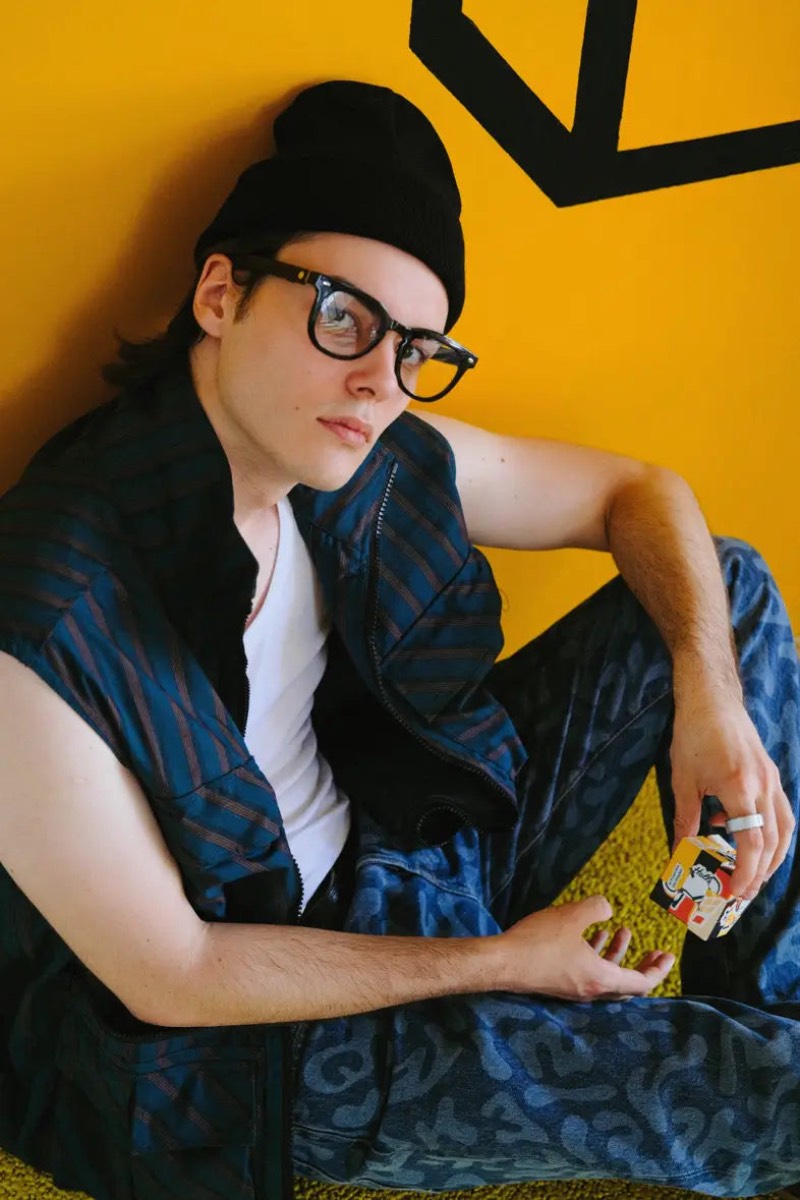 Halliday Glasses: un hombre con un gorro negro, sentado en el suelo y apoyado sobre una pared amarilla, con un estilo alternativo, lleva puestas las Halliday Glasses y un anillo con el que las controla.