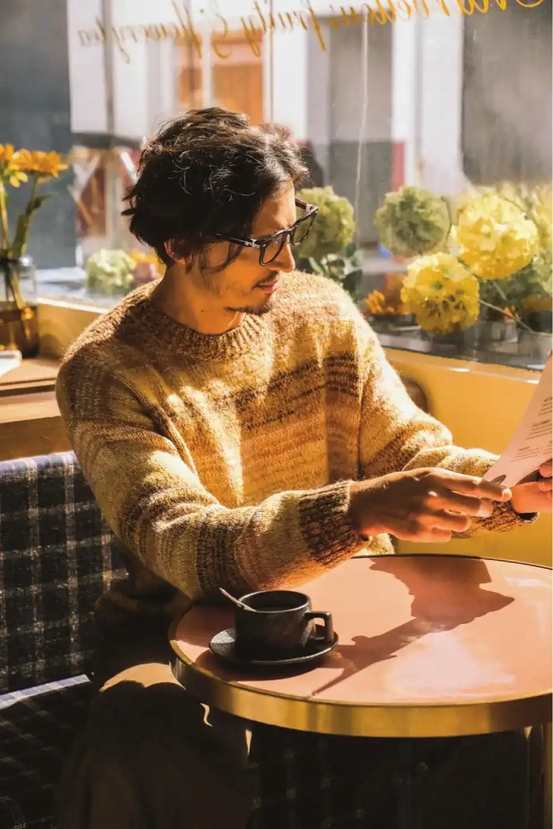 Halliday Glasses: un hombre con el pelo corto, sentado en una cafetería junto a la ventana en un día soleado con un jerse en colores tierra a rayas, lleva puestas las Halliday Glasses y lee la carta del establecimiento.