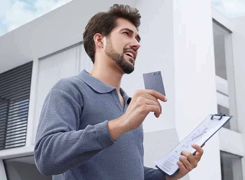 Plaud Note: foto de cuerpo medios de un chico de piel clara con suéter gris de manga larga mientras anda por la zona exterior de un edificio con una grabadora Plaud Note en una mano y una carpeta en la otra.