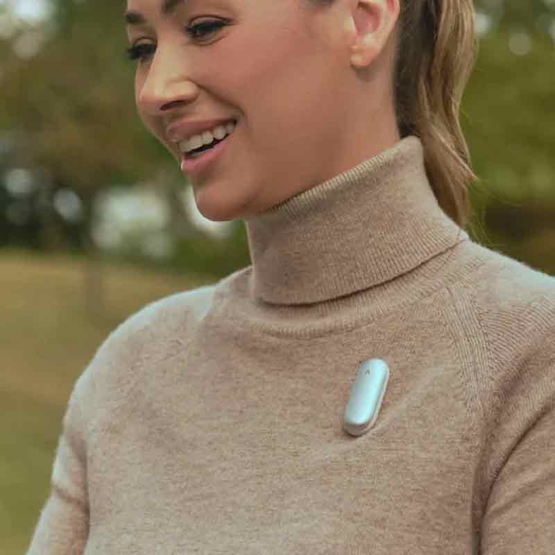 Plaud Note: primer plano de una chica sonriente de piel clara con suéter de cuello alto en color tierra en el que lleva la grabadora Plaud NotePin.