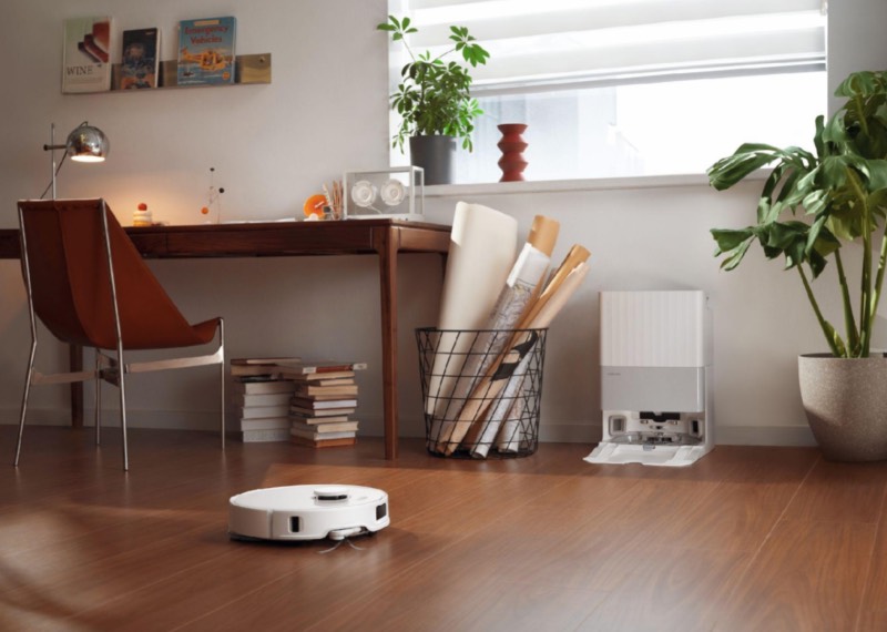 Roborock: un robot de limpieza Roborock en el suelo de madera de una habitación que tiene una mesa de escritorio con una silla, algunos libros y plantas.