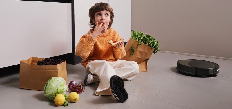 Roborock: un niño se come una tostada junto a las bolsas de la compra sentado en el suelo y cerca de un aspirador negro redondo de Roborock.