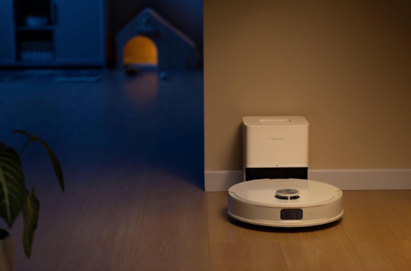 Roborock: un robot de limpieza Roborock blanco en el suelo junto a su base en un pasillo con poca luz.