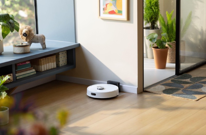 Roborock: un robot de limpieza Roborock blanco en el suelo de madera de un salón luminoso con libros y plantas.