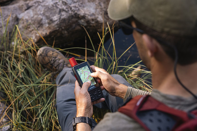 GpsMap H1i Plus: foto hecha desde arriba de un hombre sentado en una roca con mochila, gorra y gafas de sol consultando la pantalla de un dispositivo GpsMap H1i Plus en la que apetece una ruta de montaña.