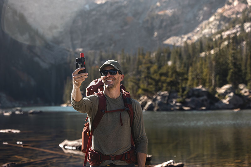GpsMap H1i Plus: un hombre con mochila granate, gorra oscura, camiseta gris y gafas de sol negras se hace un selfie con un dispositivo GpsMap H1i Plus en un día soleado en mitad de un paisaje de montaña.
