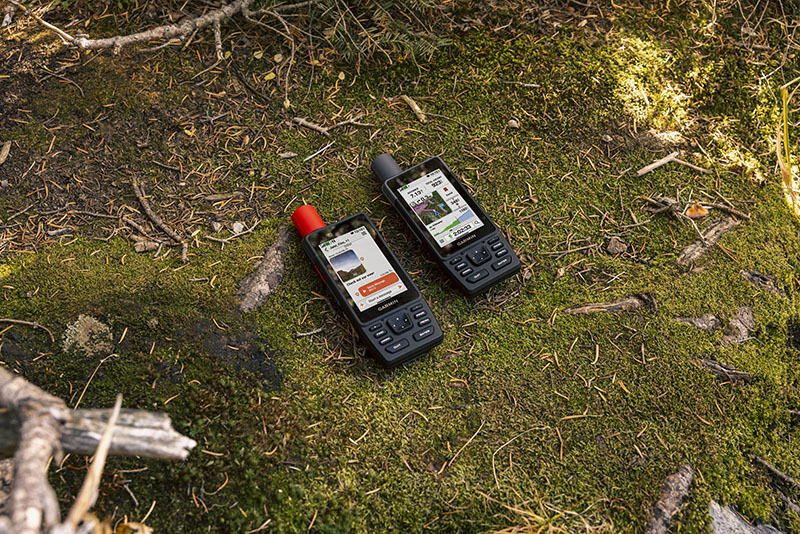 GpsMap H1i Plus: foto hecha desde arriba de dos dispositivos GpsMap H1i Plus encendidos, sobre un tramo de césped, que muestran en sus pantallas una conversación uno y datos de la ruta de montaña el otro.