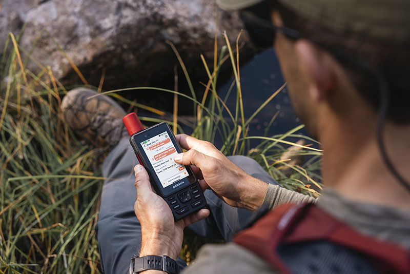 GpsMap H1i Plus: foto hecha desde arriba de un hombre sentado en una roca con mochila, gorra y gafas de sol mirando la pantalla de un dispositivo GpsMap H1i Plus en la que apetece una conversación con mensajes de texto.