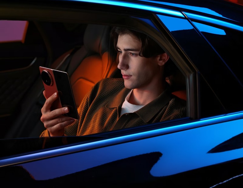 Honor Magic V5: un chico mira su teléfono Honor Magic V5 desplegado desde su asiento de un coche azul por fuera con tapicería en rojo.