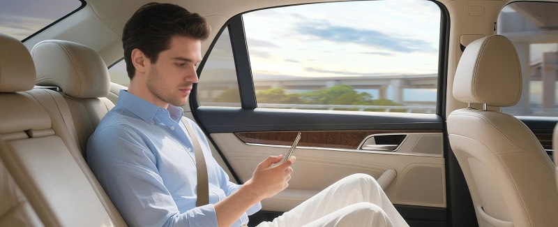 Honor Magic V5: un chico de tez blanca y pelo corto oscuro, con camisa azul y pantalón blanco, mira su teléfono Honor Magic V5 en la parte trasera de un coche en movimiento que tiene asientos de piel tapizados en beige.