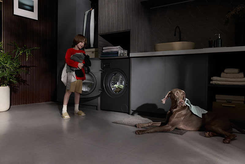 lavadora-secadora Roborock Zeo X: un niño con un montón de ropa entre sus brazos junto a la lavadora-secadora Roborock Zeo X abierta, integrada en un mueble moderno, y junto a un perro grande oscuro tumbado en el suelo.