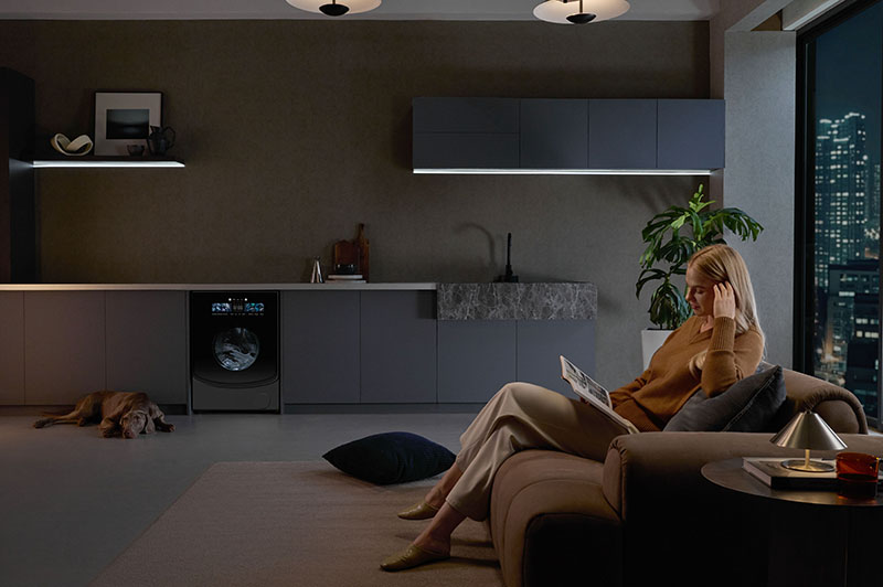 lavadora-secadora Roborock Zeo X: lavadora-secadora Roborock Zeo X integrada en un mueble de cocina de un espacio interior moderno y minimalista que se junta con el salón en el que hay una chica rubia leyendo en el sofá y un perro grande tumbado en el suelo.