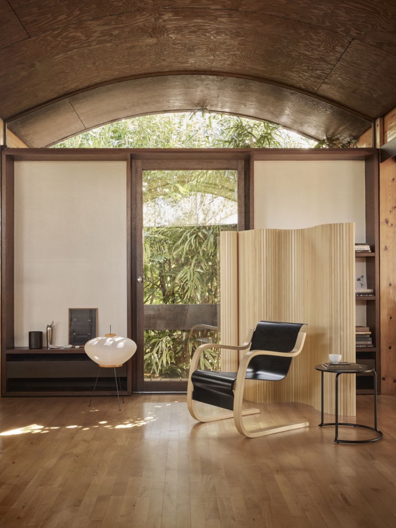 Artek: salón amplio y luminoso, con frondosa vegetación tras el cristal, que incluye una silla delante de un biombo de madera y junto a una mesa auxiliar.