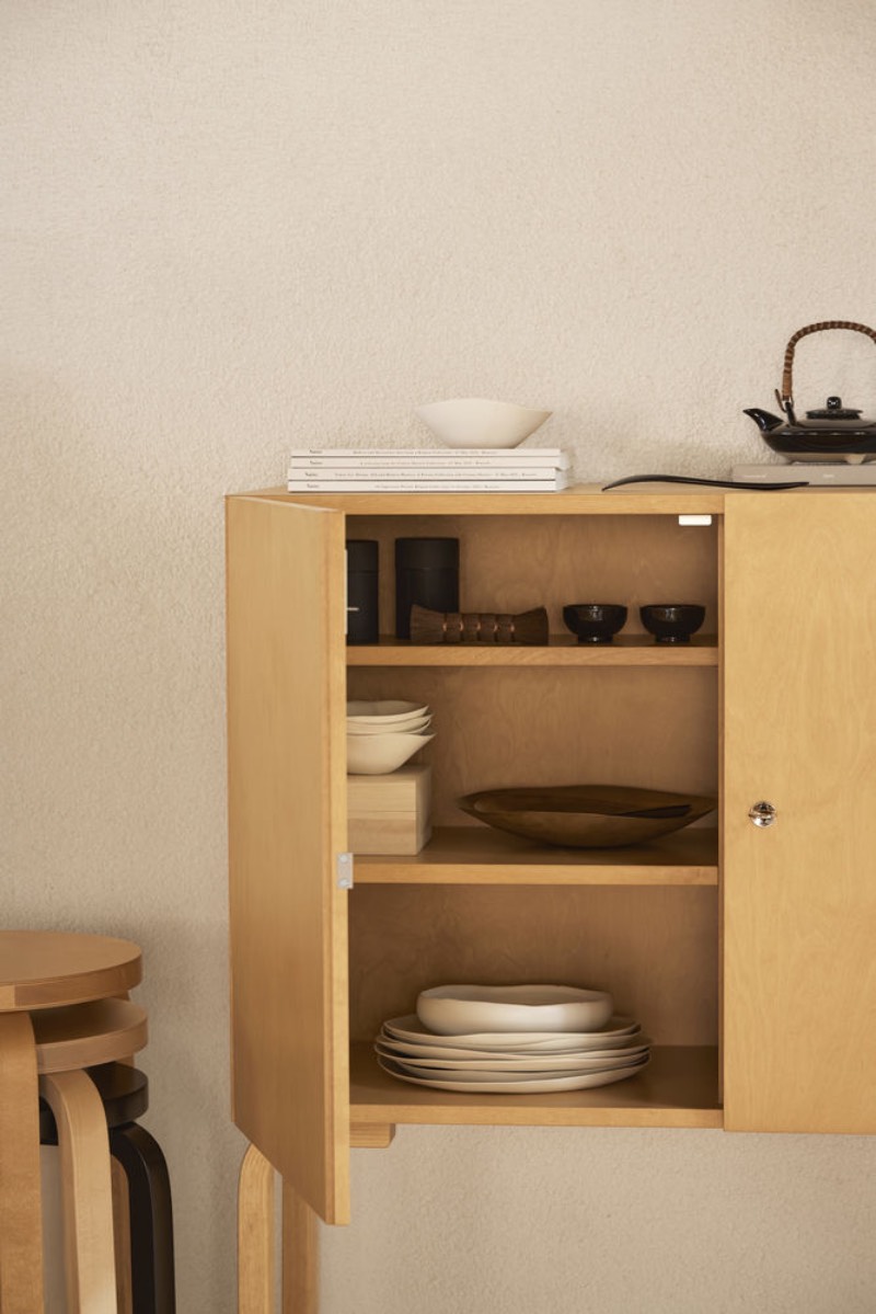 Artek: Cabinet 250 de la colección Gems from the Archive de Artek con una puerta abierta, vajilla en su interior y junto a unos taburetes en el mismo tono de madera.