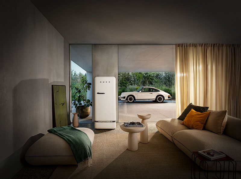Porsche x Smeg: una zona de estar con un sofá grande en beige y dos mesas tipo setas en el mismo tono, en un espacio con amplios cristales que dan a un jardín en el que está aparcado un coche blanco de Porsche y en un trozo de pared hay un frigorífico blanco de la colección de Porsche x Smeg.