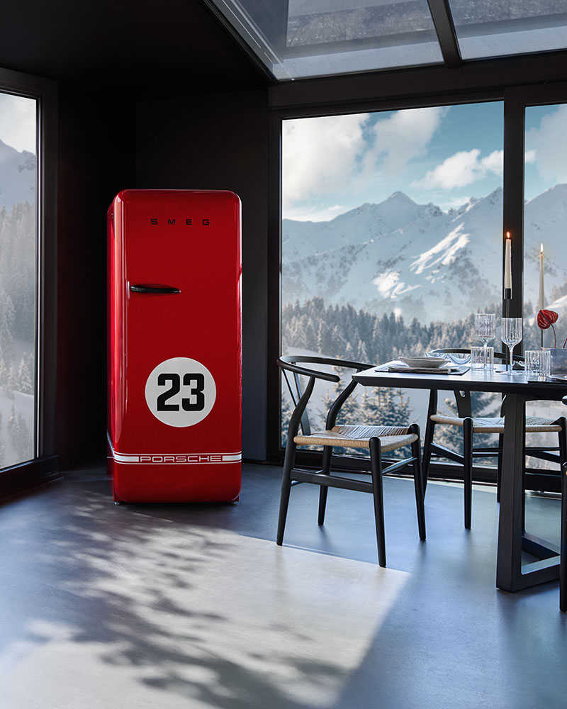 Porsche x Smeg: frigorífico rojo de la Colección 917 Salzburg de Porsche x Smeg en una esquina de un comedor luminoso, con amplios ventanales que dan a una zona de montañas nevadas.