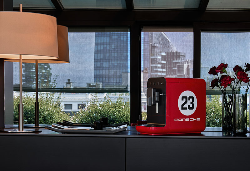 Porsche x Smeg: máquina de café en rojo de la Colección 917 Salzburg de Porsche x Smeg sobre una superficie con una lámpara, una bandeja con tazas en negro y un jarrón con rosas rojas, que da a un ventanal de ciudad con edificios altos.