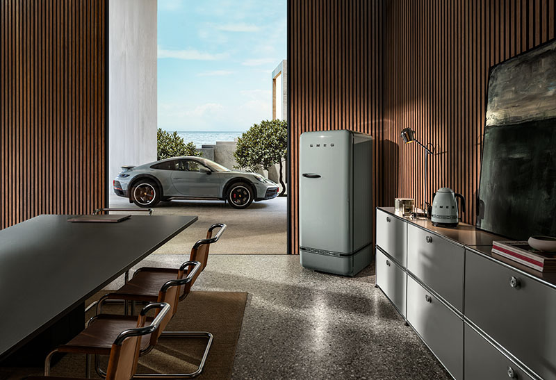 Porsche x Smeg: una zona tipo comedor o sala de reuniones en tonos grises con madera, que cuenta con un frigorífico gris de la colección de Porsche x Smeg en una esquina, y todo en un espacio con una puerta muy grande que da a una zona exterior privada en la que está aparcado un coche gris de Porsche.