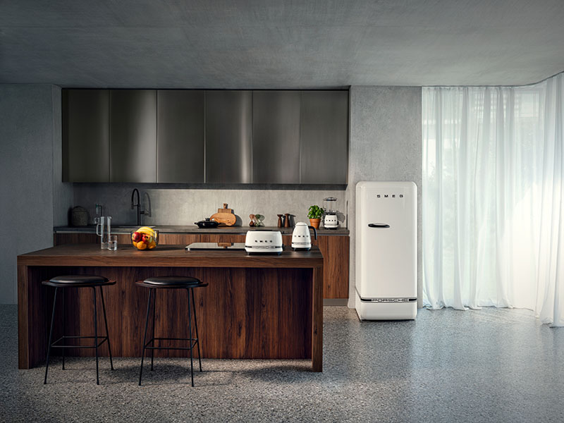 Porsche x Smeg: amplia cocina luminosa con suelo, paredes y techos en gris, muebles en madera oscura, pequeños electrodomésticos de la colección de Porsche x Smeg en la encimera y un frigorífico blanco a juego en un trozo de pared junto a las cortinas traslúcidas blancas.
