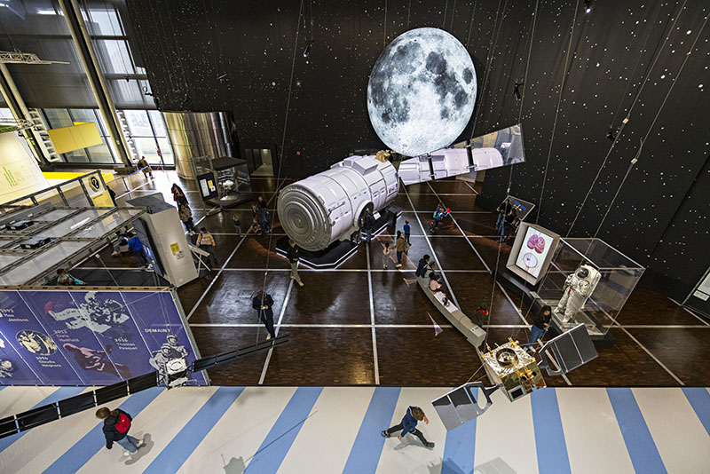 Vista general de la exposición Space Mission en la Cité des Sciences et de l’Industrie, con una gran Luna suspendida y el módulo I-Hab en el centro de la sala