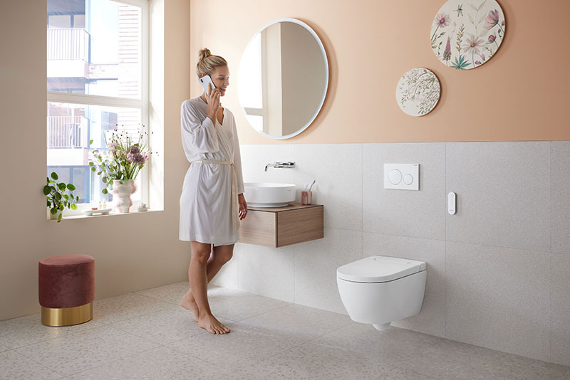 Inodoro bidé Geberit AquaClean: una mujer de pelo rubio con un moño y bata blanca de seda en un baño amplio en tonos claros hablando por teléfono junto a la pared del espejo, lavabo e Inodoro bidé Geberit AquaClean.