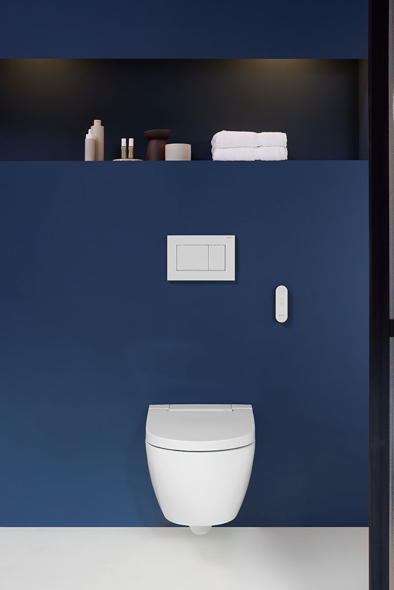 Inodoro bidé Geberit AquaClean: el Inodoro bidé Geberit AquaClean suspendido en una pared en color azul oscuro con una balda superior que tiene un par de toallas y otros productos de baño.
