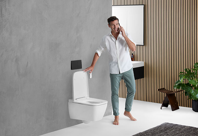 Inodoro bidé Geberit AquaClean: un hombre de pelo castaño con un pantalón vaquero verde y una camisa blanca descalzo en un baño amplio hablando por teléfono se dispone a bajar la tapa de un Inodoro bidé Geberit AquaClean suspendido en una pared gris degradada.